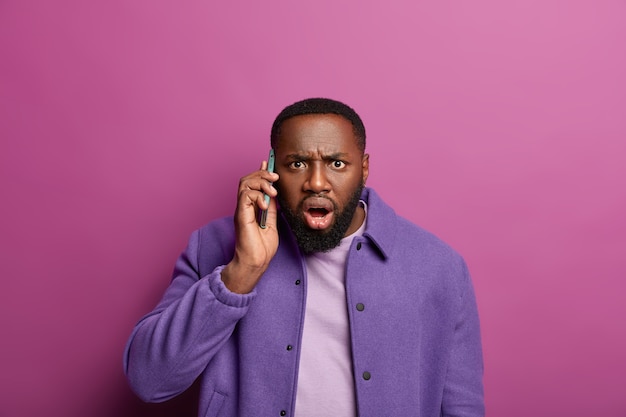 Shocked black man stares in panic, learns bad news during telephone call, gasps from wonder, frowns face and looks nervously, wears purple jacket