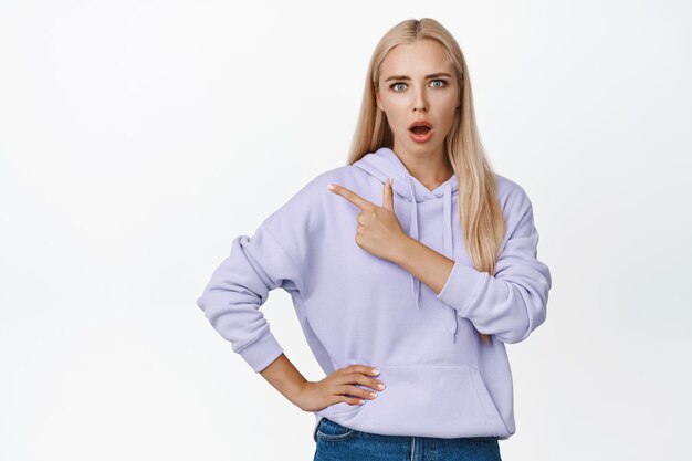 Shocked blond girl pointing finger left looking worried at camera standing concerned against white background copy space