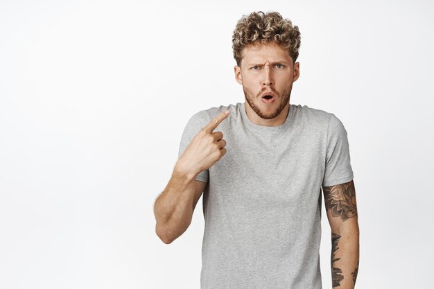 Shocked blond guy gasps offended pointing finger at himself and staring insulted at camera standing in gray tshirt over white background