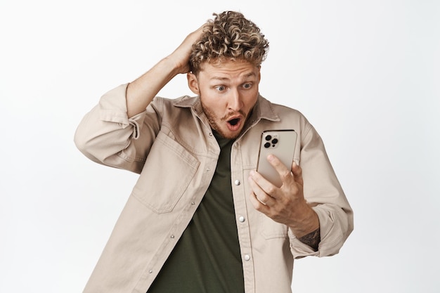 Шокированный блондин смотрит на мобильный телефон с испуганным выражением лица, задыхаясь от трепета, стоя на белом фоне
