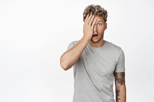 Shocked blond man facepalm staring in awe at camera drop jaw and looking in disbelief at advertisement standing in gray tshirt over white background