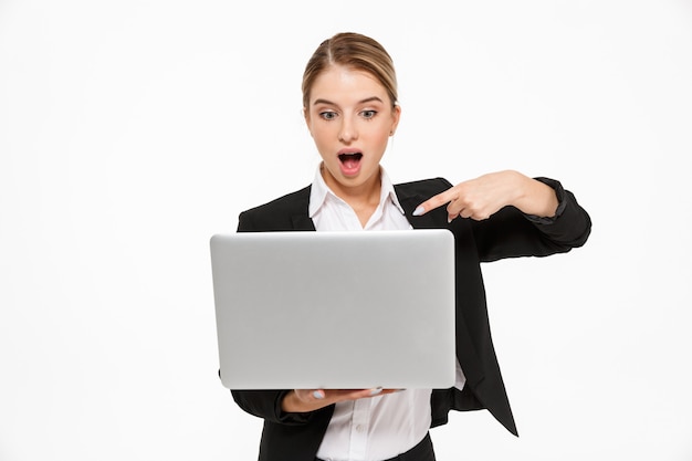 Shocked blonde business woman holding laptop computer and pointing at him over white wall