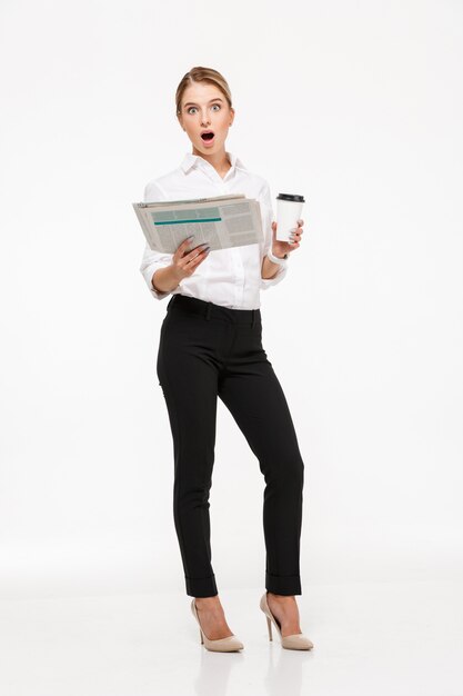 Shocked blonde business woman holding newspaper and cup of coffee while  over white wall