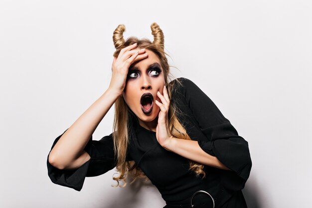 Shocked blonde woman in halloween costume standing . Scared witch posing on white wall.