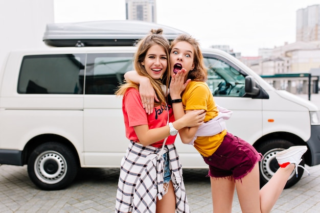 Free photo shocked brunette girl in yellow shirt standing on one leg and embrace her lovely sister in pink attire