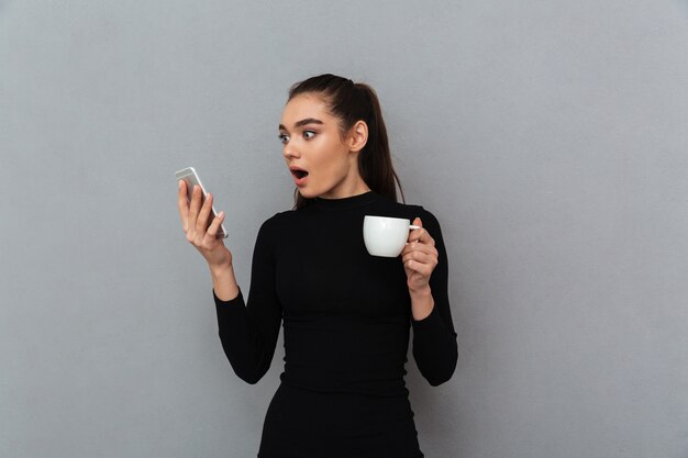 Shocked brunette woman in black clothes looking at smartphone