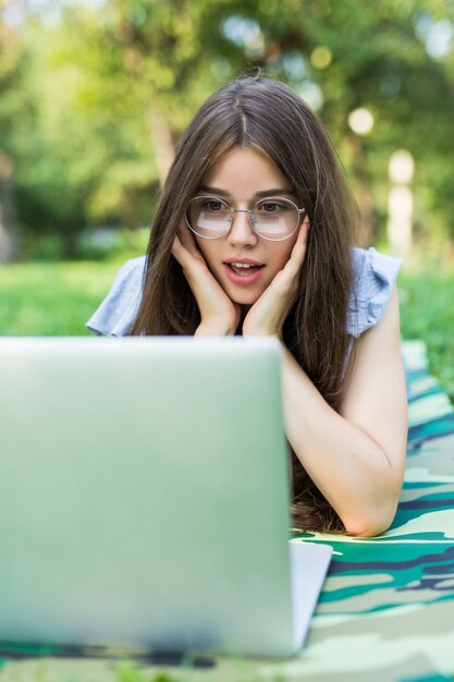 Шокированная брюнетка женщина в очках, лежа на траве в парке и с помощью портативного компьютера
