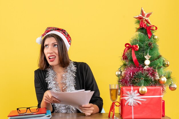 산타 클로스 모자와 새 해 장식 문서를 들고 사무실에서 크리스마스 트리가있는 테이블에 앉아 양복에 충격을받은 비즈니스 아가씨