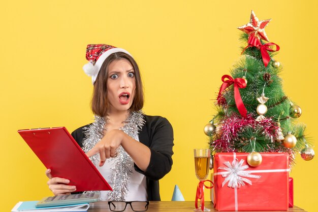 Shocked business lady in suit with santa claus hat and new year decorations looking at document and sitting at a table with a xsmas tree on it in the office