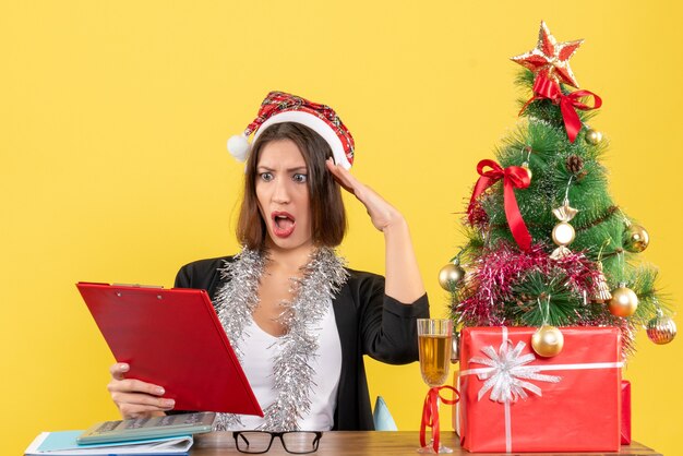 Shocked business lady in suit with santa claus hat and new year decorations looking at document and sitting at a table with a xsmas tree on it in the office