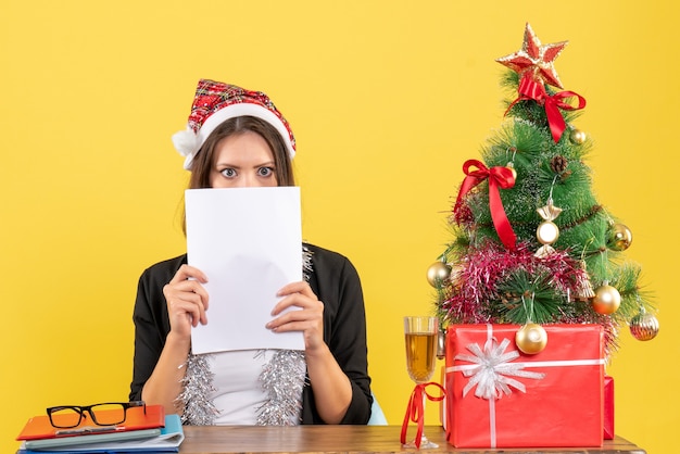 Shocked business lady in suit with santa claus hat and new year decorations working alone and sitting at a table with a xsmas tree on it in the office