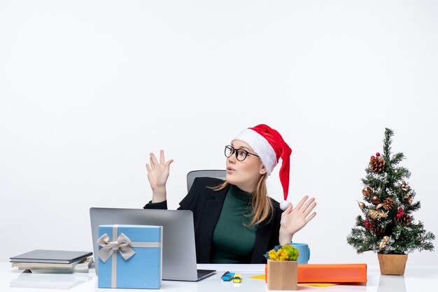 Donna d'affari scioccata con cappello di babbo natale seduto a un tavolo con un albero di natale e un regalo su di esso che punta sopra su sfondo bianco