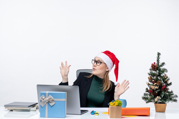 Шокированная деловая женщина в шляпе санта-клауса сидит за столом с рождественским деревом и подарком на нем, указывая выше на белом фоне