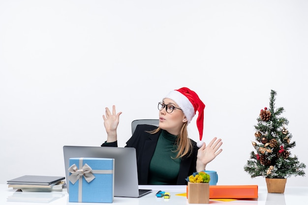 Shocked business woman with santa claus hat sitting at a table with a Xsmas tree and a gift on it pointing above on white background