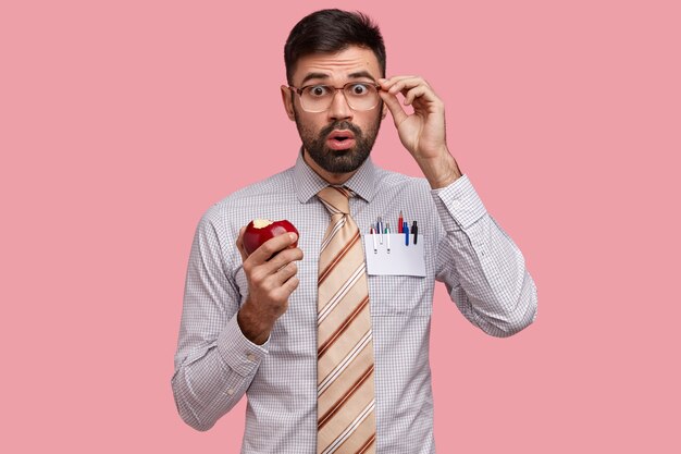 Шокированный бизнесмен, одетый в строгую рубашку и галстук, ест вкусное яблоко, недоуменно смотрит в очки