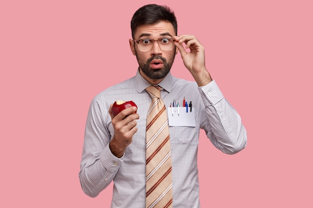 Shocked businessman dressed in formal shirt and tie, eats delicious apple, looks through spectacles in bewilderment