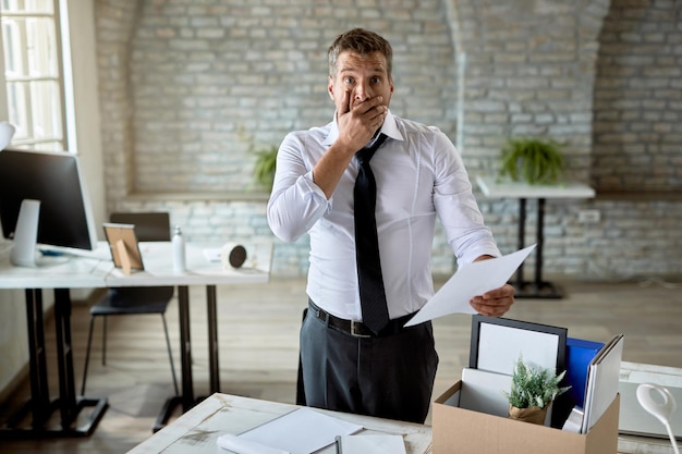 Free photo shocked businessman standing in the office and reading a notice after getting fired from his job