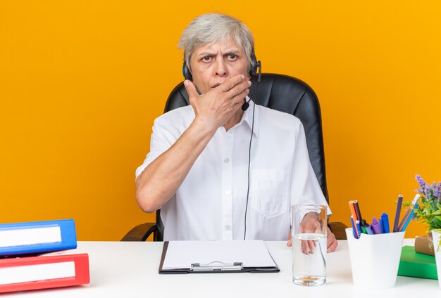 Shocked caucasian female call center operator on headphones sitting at desk with office tools putting hand on mouth