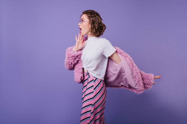 Free photo shocked caucasian girl in white t-shirt and fur jacket posing on purple wall. indoor portrait of emotional woman with wavy hairstyle having fun