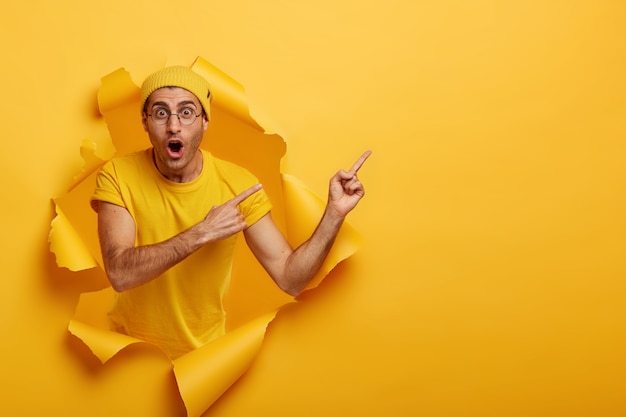 Shocked Caucasian man keeps mouth widely opened mouth, points index fingers aside on blank space, stares through round glasses, stands in paper hole