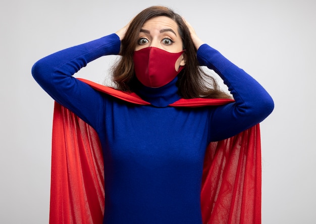 Free photo shocked caucasian superhero girl with red cape wearing red protective mask puts hands on head and looks at camera