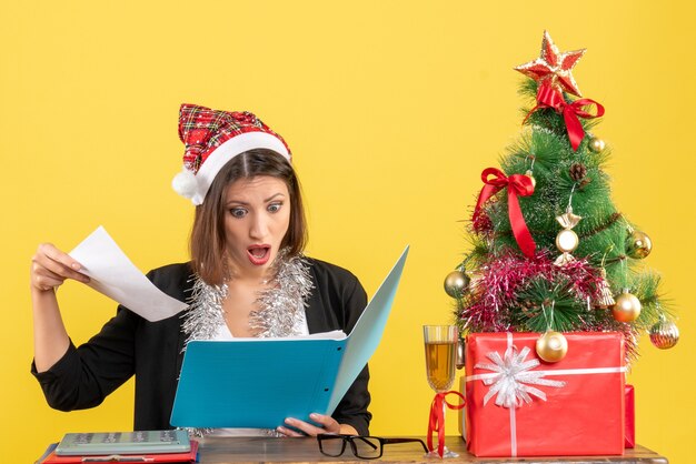 Shocked charming lady in suit with santa claus hat and new year decorations holding document in the office on yellow isolated 