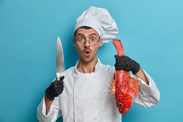 Shocked chef stares with great surprisement, holds sharp knife, whole fresh fish, prepares healthy food quickly, sea bass soup