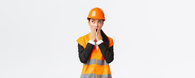 Shocked and concerned asian female engineer architect in safety helmet and reflective jacket looking upper left corner and gasping worried stare amazed standing white background