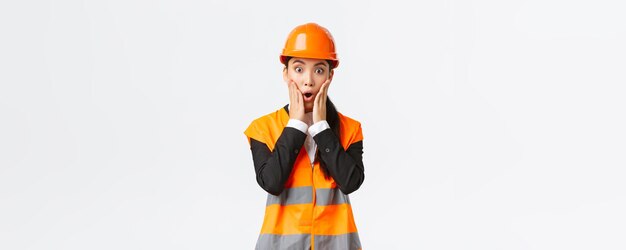 Shocked and concerned asian female engineer having problem at building area stare at project astounded with panic face holding hands near mouth and gasping worried standing white background