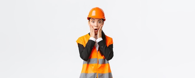 Шокированная и обеспокоенная азиатская женщина-инженер, имеющая проблемы на строительной площадке, смотрит на проект, пораженная паническим лицом, держась за руки возле рта и задыхаясь от беспокойства на белом фоне