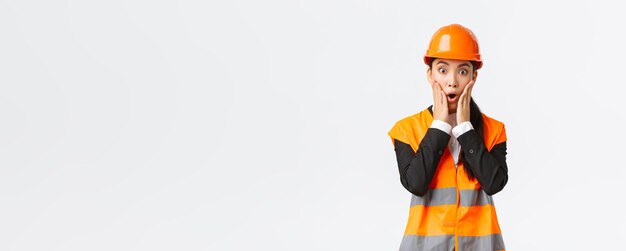 Shocked and concerned asian female engineer having problem at building area stare at project astounded with panic face holding hands near mouth and gasping worried standing white background