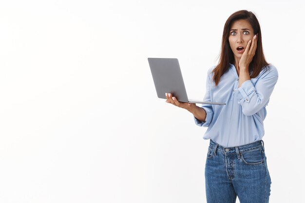 Shocked concerned brunette adult woman with tattoo, panic, stare distressed worried at front, touch cheek ambushed, gasping anxious, hold laptop, delete file, receive bad news text message