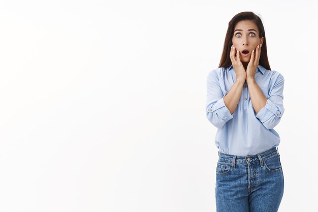 Shocked concerned brunette woman feel scared ambushed horrible news, gasping frightened, touch face, stare at front terrified, drop jaw astonished, panicking, stand white wall