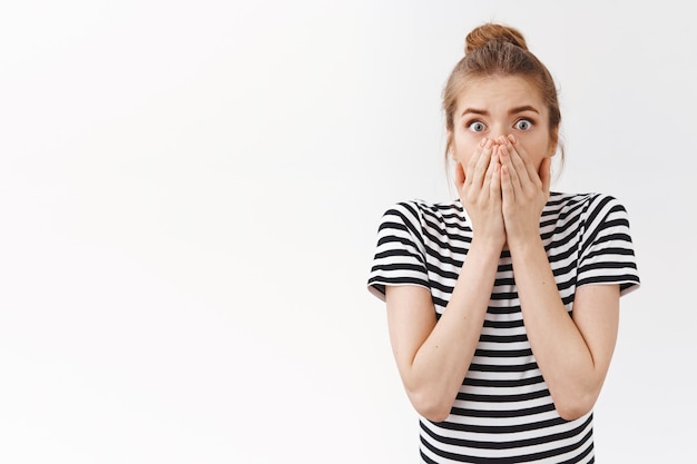 Shocked concerned young caucasian woman with messy bun, gasping holding breath stunned, cover mouth with hands and stare camera scared for you, frightened caused accident, white background