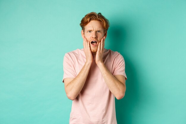 Шокированный и обеспокоенный молодой человек с рыжими волосами, смотрящий в камеру с обеспокоенным и трогательным лицом, стоит в футболке на бирюзовом фоне.