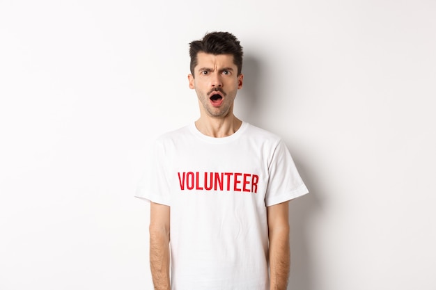 カメラを見つめ、白い背景に立って不機嫌に眉をひそめているボランティアのTシャツでショックを受けて混乱した男