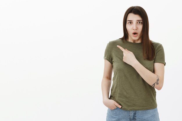 Shocked and confused nervous girl gasping open mouth in frustration and pointing at upper left corner questioned, asking what happened with worried and anxious expression, concerned over gray wall