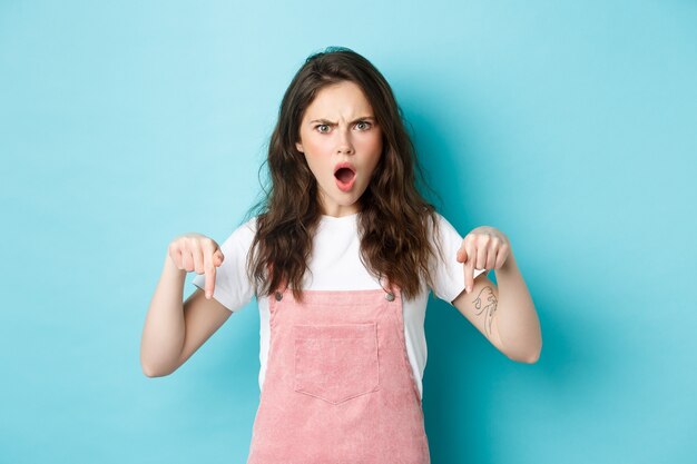 Shocked and confused young woman gasping and looking startled at camera, pointing fingers down, standing in summer clothes against blue background