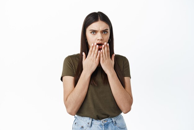 Shocked and confused young woman open mouth and frowning displeased staring startled at something shocking bad standing against white background