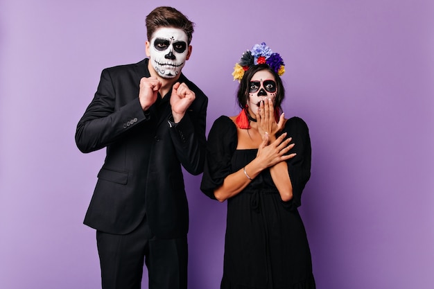 Shocked couple with skull-shaped masks in frightened posing on purple wall. Portrait of guy in black suit and girl in dark dress with bright accents.