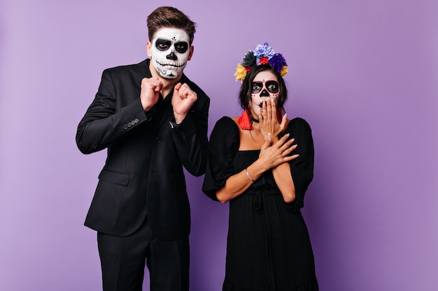 Бесплатное фото Шокированная пара с масками в форме черепа в испуганном позировании на фиолетовой стене. портрет парня в черном костюме и девушки в темном платье с яркими акцентами.
