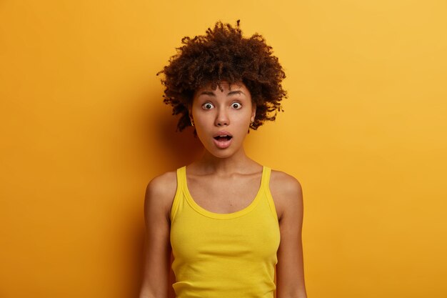 Shocked curly ethnic woman looks anxiously and worried, notices how accident happened, opens mouth with bewilderment, wears casual clothes, has widely opened eyes, isolated on yellow wall