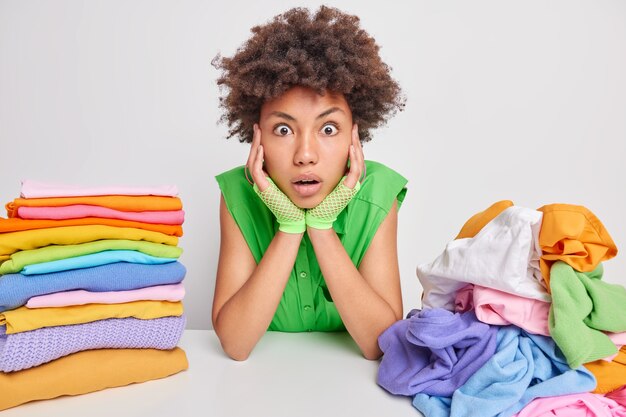 Shocked curly haired Afro American housewife stares surprisingly keeps hands on cheeks cannot believe her eyes has much housework folds washed laundry isolated over white