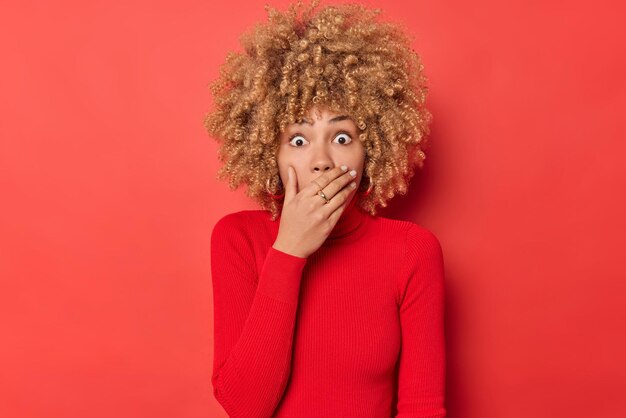 Shocked curly haired woman gasps from wonder covers mouth with hand stares bugged eyes witnesses terrible scene wears casual turtleneck isolated over red background. Human reactions concept.