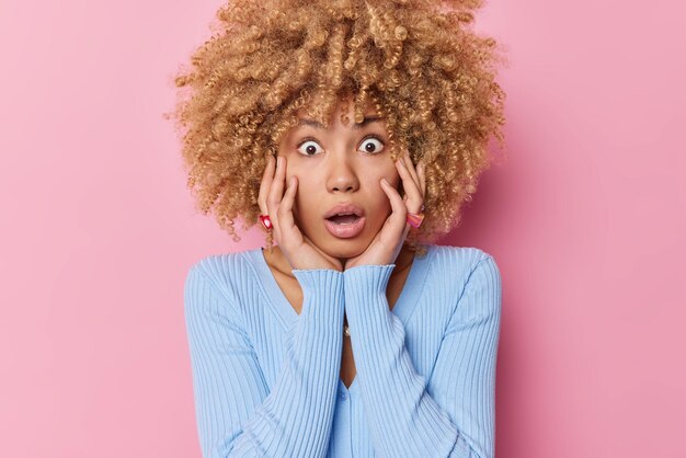 Shocked curly haired woman keeps hands on cheeks stares bugged eyes at camera has surprised exression holds breath wears casual blue jumper isolated over pink background Human reactions concept