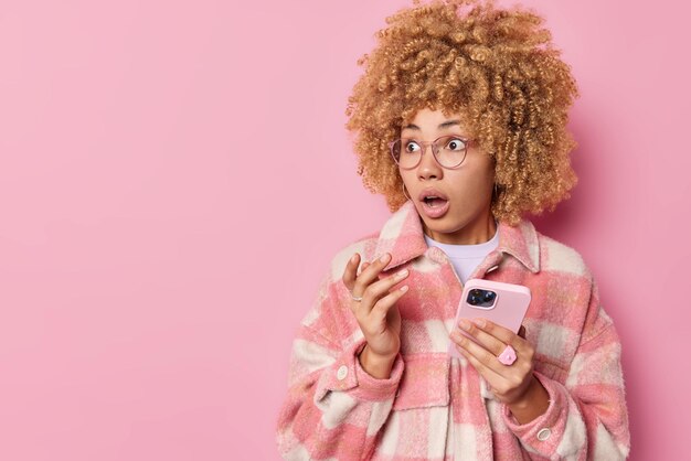 Шокированная кудрявая женщина выглядит с испуганным испуганным выражением лица, держит рот открытым, держит смартфон, одетый в модную клетчатую куртку, изолированную на розовом фоне пустое место для текста