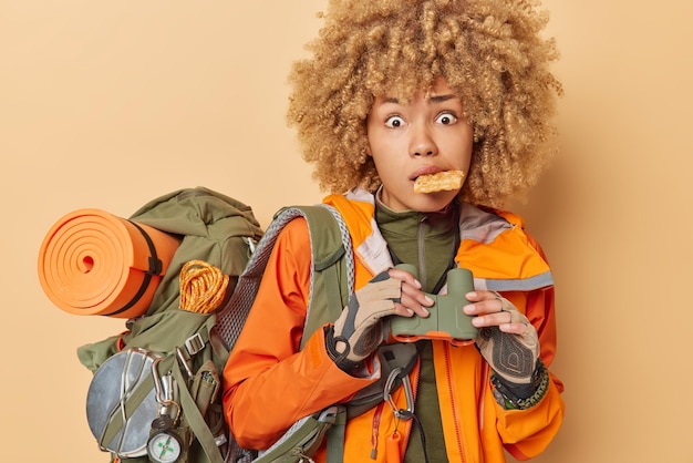 Free photo shocked curly haired woman spends vacation actively keeps waffle in mouth holds binoculars carries heavy rucksack dressed in casual clothes isolated over brown background tourist adventure