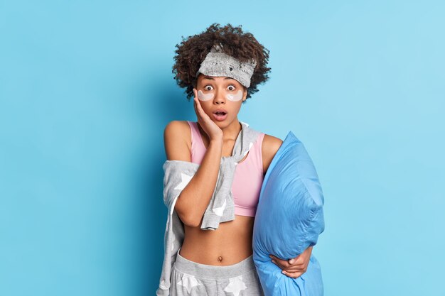 Shocked curly haired woman stares at camera with stunned expression keeps hand on cheek holds breath from wonder dressed in nightwear isolated over blue wall