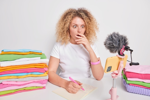 Shocked curly haired woman stares at smartphone webcam finds out amazing newsmakes notes in notepad sits at table with two stacks of folded laundry isolated over white 