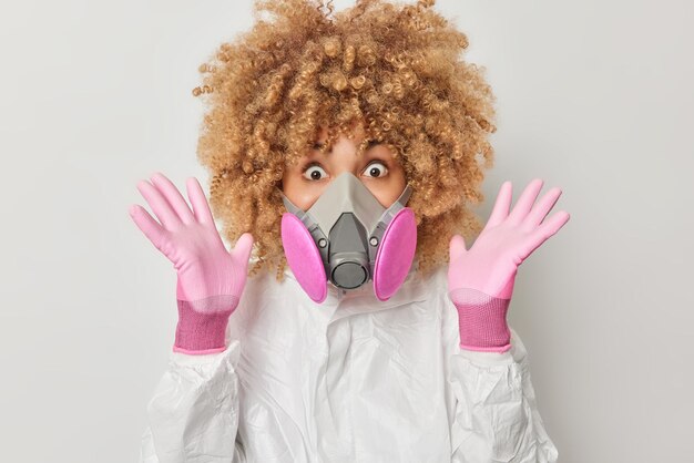 Shocked curly haired young woman keeps palms raised wears chemical protective suit rubber gloves and respirator or breathing gas mask works on virus research poses indoor against grey background